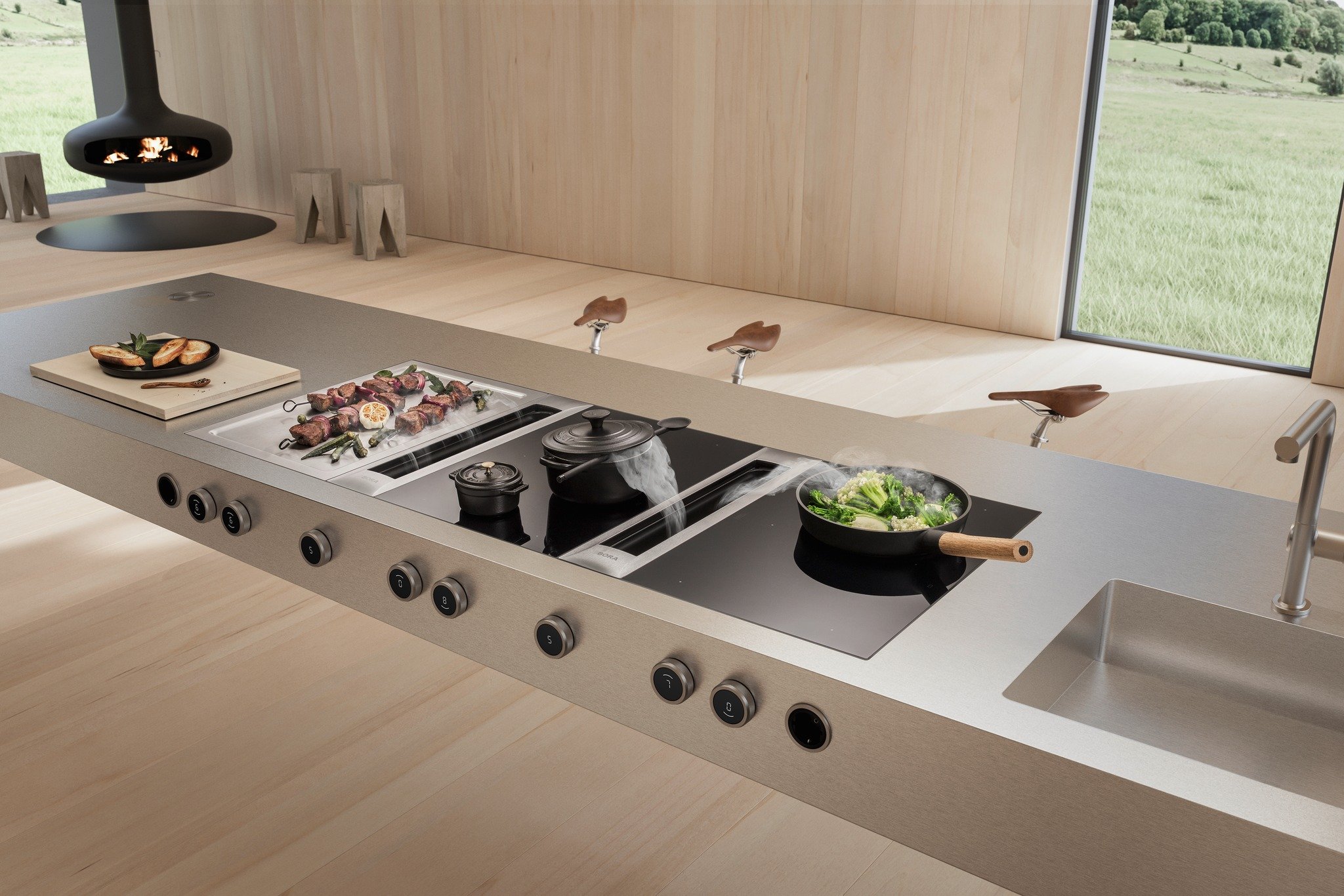 Modern kitchen island with built-in stovetop, pots steaming food, and a frying pan with vegetables, set in a light wood room with large window and hanging fireplace.